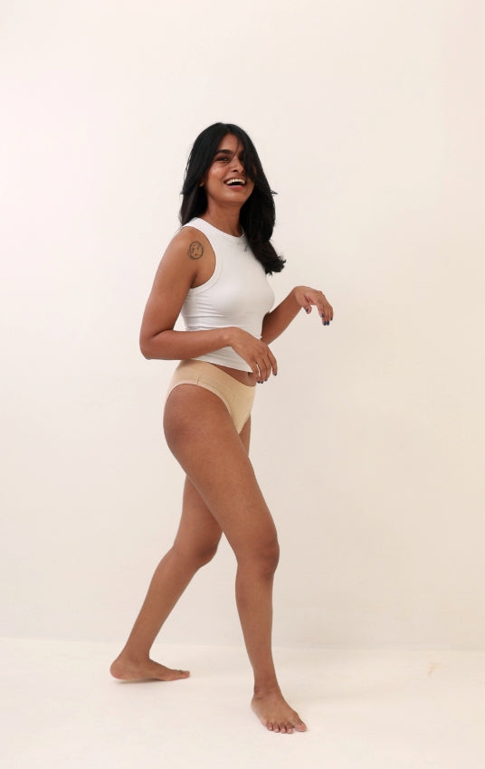 A woman modeling a white sleeveless crop top and beige high-waisted briefs against a plain off-white studio background.
