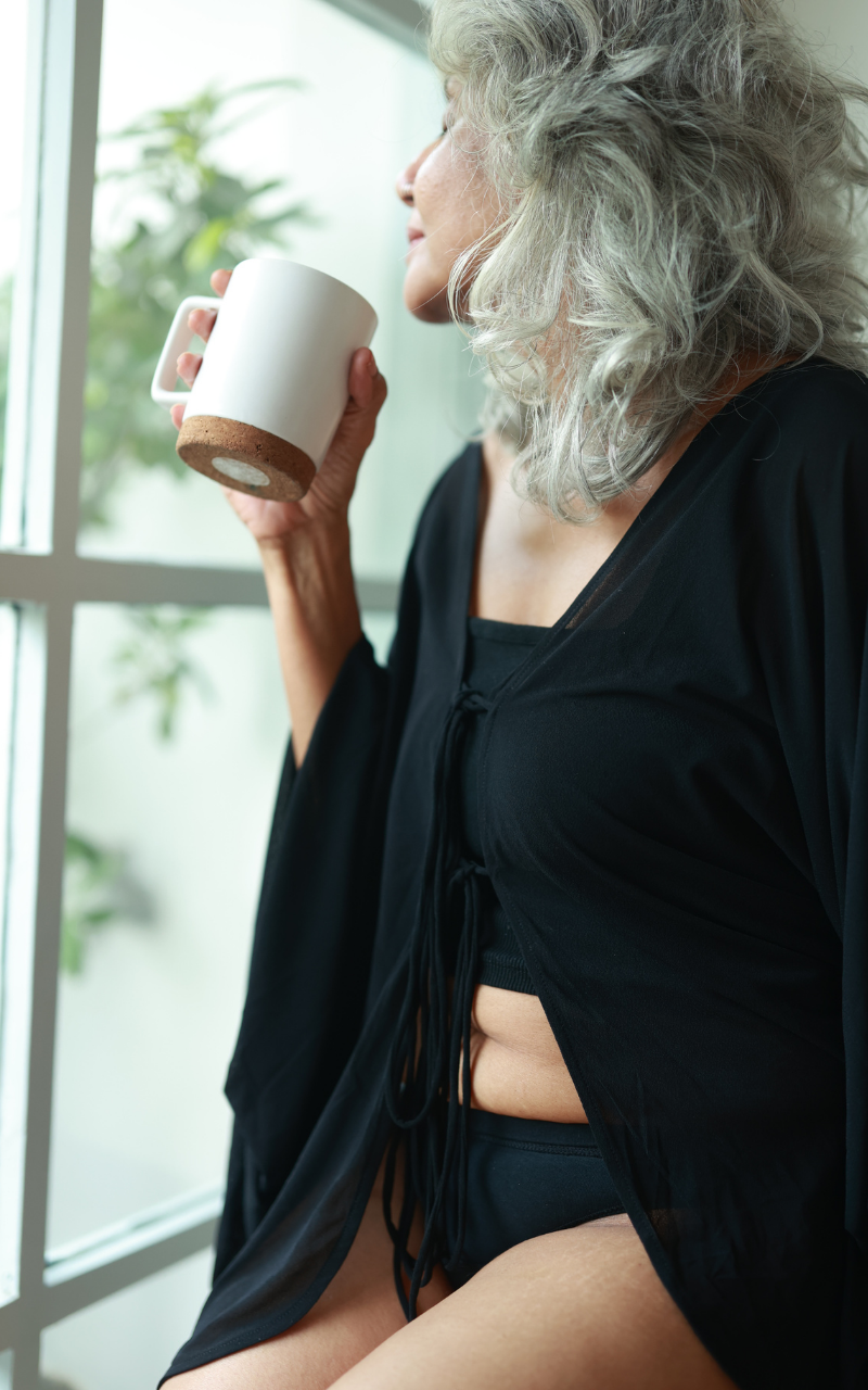 A woman with gray hair sits by a window, holding a white ceramic mug with a cork base. She is wearing a black open-front cardigan over a black top and shorts.