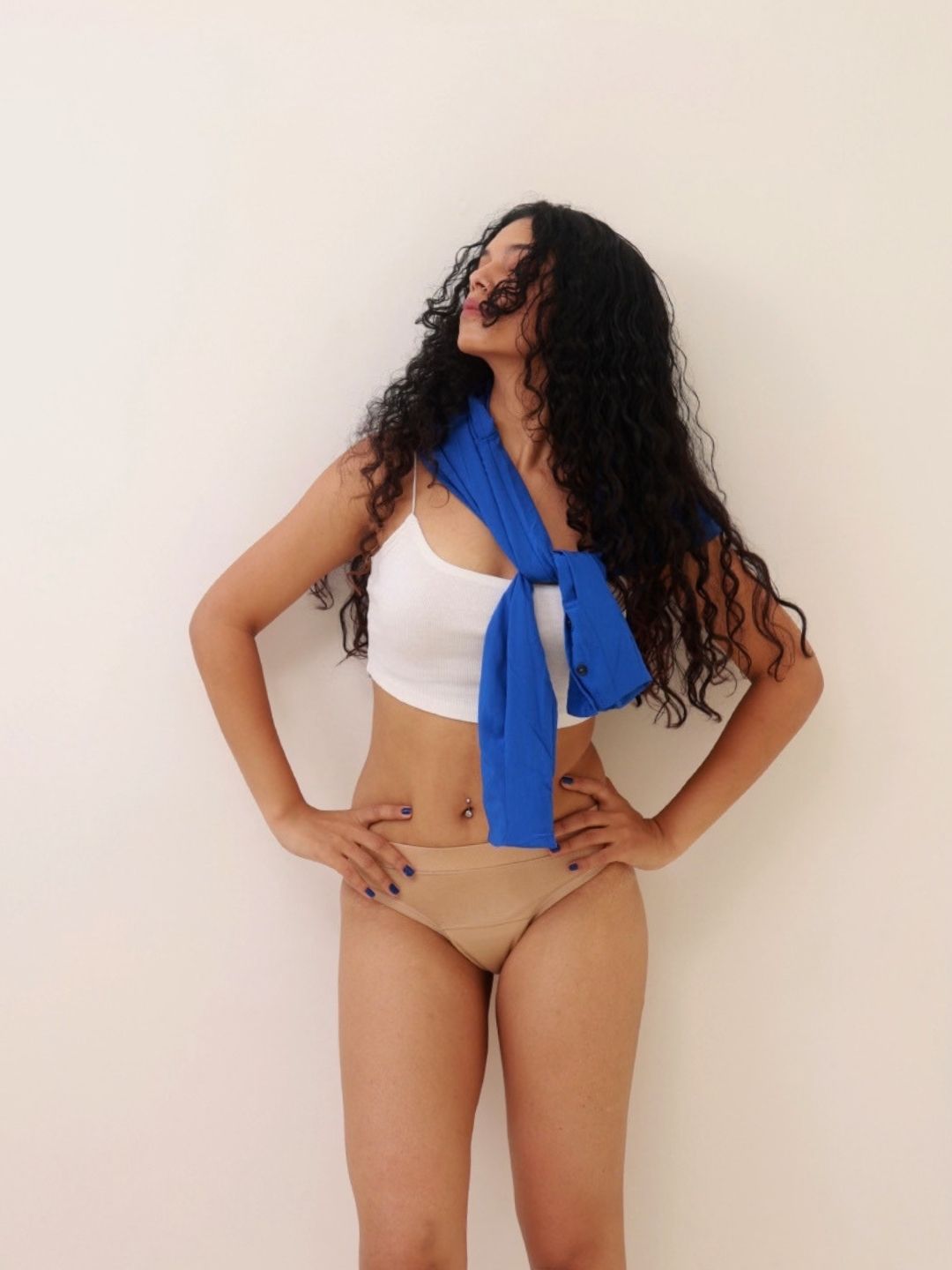 Ride or Dry leakproof underwear in Oatmeal: a woman with long curly black hair against a plain off-white wall, wearing a white crop top, beige bikini-style underwear and a bright blue shirt tied around her shoulders.