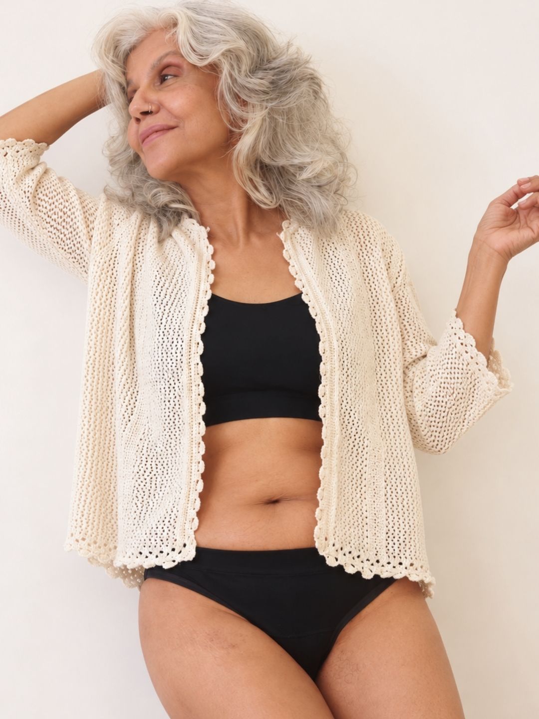 Ride or Dry leakproof underwear in Volcanic Black: a woman with gray hair wearing a cream open-front crochet cardigan over a black bra and black underwear against a plain off-white background.